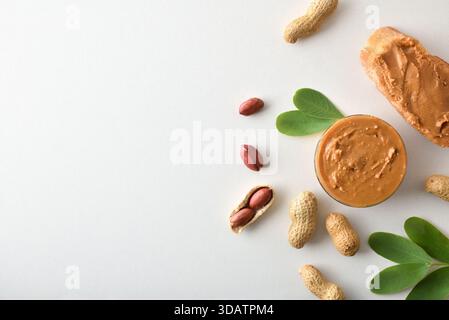 Bowl full of peanut butter and slice of bread on white table with fruits and leaves. Top view. Stock Photo