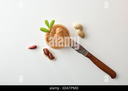 Detail of creamy peanut butter in a glass bowl with a buttered knife resting on it, isolated on a white table with nuts and peanut kernels. Top view. Stock Photo