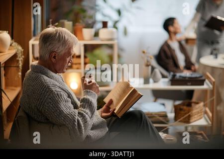 Senior Caucasian Man Reading Book and Drinking Coffee in Nursing Home Lounge Stock Photo