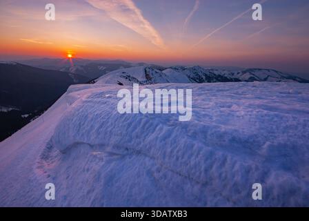 View of the sun setting behind snow-covered mountain peaks, casting an orange glow across the pristine white landscape, Rovna hola, Banskobystricky kr Stock Photo