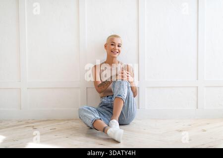 Portrait of young happy caucasian woman with short hair sitting on floor and smiling at camera, leaning on white wall Stock Photo