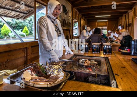 Ama diver Kimiyo Hayashi with fresh seafood, which she prepares on the charcoal grill right in front of the guests. The guests sit around the open grill, chatting directly with the Ama diver. The hut is a replica of a traditional Ama hut, Ise Bay, Shima, Mie, Japan Stock Photo