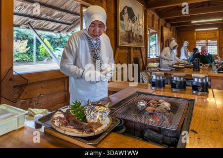 Guests sit around the open grill, chatting directly with Ama diver Kimiyo Hayashi as she prepares fresh seafood on the charcoal grill, in a replica of a traditional ama hut, Ise Bay, Shima, Mie Prefecture, Japan. Stock Photo