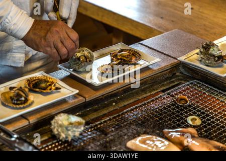 Ama diver Kimiyo Hayashi with fresh seafood, which she prepares on the charcoal grill right in front of the guests. The guests sit around the open grill, chatting directly with the Ama diver. The hut is a replica of a traditional Ama hut, Ise Bay, Shima, Mie, Japan Stock Photo