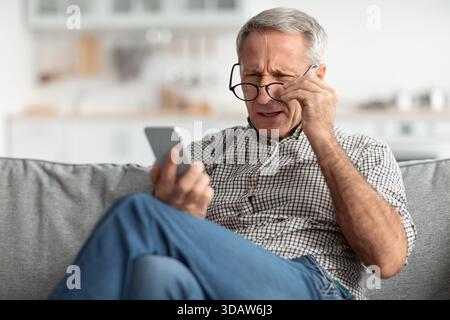 Older Man Sitting on Sofa Looking at Phone With Concentration and Concern in a Modern Living Room Stock Photo