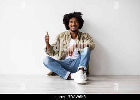 Cheerful Black Millennial Guy Giving Thumbs up While Relaxing Indoors Near White Wall Stock Photo