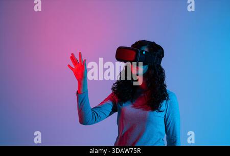 Woman in VR headset illuminated by blue and red neon lights interacting with virtual digital interface gesture Stock Photo