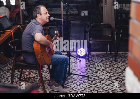 A male musician passionately performs at a live concert, seated on a chair, playing an acoustic guitar and singing into a microphone on a patterned fl Stock Photo