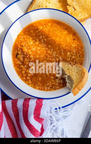 A hearty bowl of traditional Greek lentil soup, known as fakes, served with a slice of fresh bread. The soup is on a white table, accompanied by a red Stock Photo
