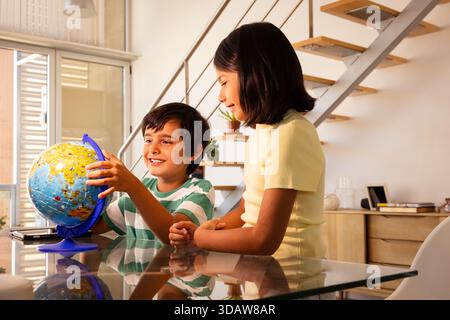 Two children happily explore a colorful globe on a glass table in a modern home setting, showcasing curiosity and learning in a bright and inviting en Stock Photo