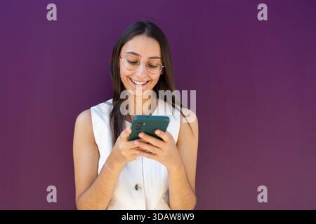 Confident businesswoman standing against a bright purple wall, smiling while using her smartphone, illustrating modern work connectivity and communica Stock Photo