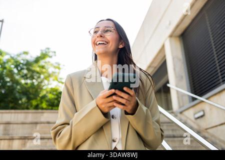 A confident businesswoman holds a smartphone, smiling brightly while outdoors. She is dressed in formal attire, representing success and positivity in Stock Photo