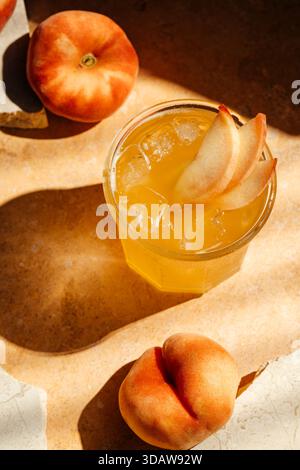 Top view of a refreshing peach drink with ice cubes in a glass, garnished with peach slices. Surrounded by whole peaches on a warm, textured stone sur Stock Photo