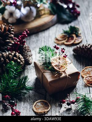 A beautifully wrapped Christmas gift adorned with pine sprigs and a dried orange slice, set amidst pine cones and berries on a rustic wooden table for Stock Photo