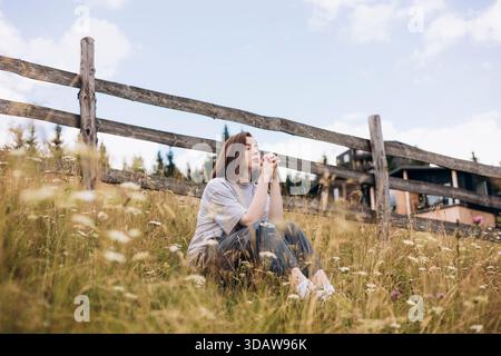 A young woman sitting peacefully amidst the grassy fields of a mountainous landscape. The serene environment and clear blue sky highlight the tranquil Stock Photo