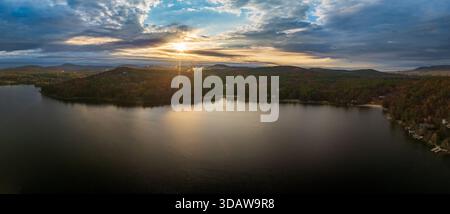 Aerial view of the sun's golden rays piercing through the clouds and reflecting on the tranquil waters of the lake surrounded by forests, Alton, New H Stock Photo