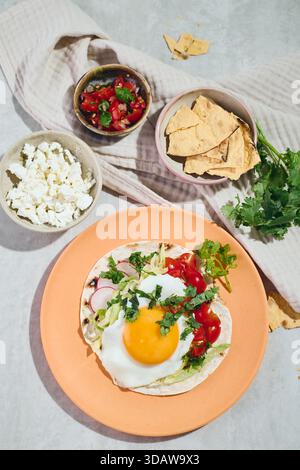 Top view of rancheros eggs on a tortilla, featuring a sunny-side-up egg, salsa, and fresh cilantro. Surrounded by feta, salsa, and chips, on a rustic Stock Photo