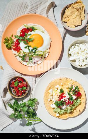 Top view of rancheros eggs on soft tortillas, garnished with fresh cilantro, tomatoes, and cheese. Served on vibrant orange and white plates with side Stock Photo