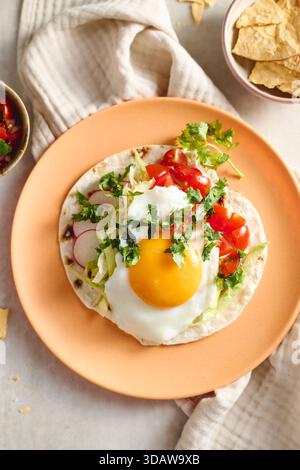 Top view of vibrant huevos rancheros featuring a fried egg on a tortilla, garnished with fresh tomatoes, radishes, and cilantro on a rustic orange pla Stock Photo
