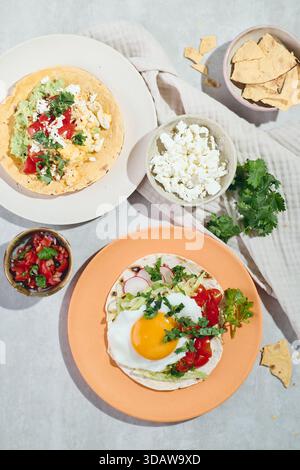 Top view of huevos rancheros with fried eggs, avocado, cheese, and tomatoes on tortillas. Served with salsa and garnished with cilantro on a soft beig Stock Photo