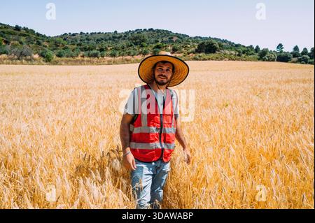 A farmer stands amidst a golden barley field in Guadalajara, Spain. Dressed in a red vest and straw hat, he embodies the essence of traditional Spanis Stock Photo