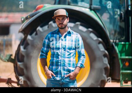 A farmer standing confidently in front of a large green tractor in the agricultural fields of Guadalajara, Spain, showcasing rural life and farming eq Stock Photo
