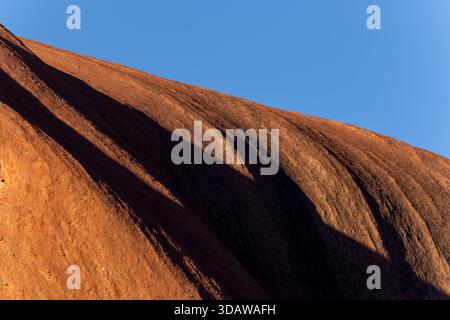 Uluru basks in the warm hues of the setting sun, highlighting its textured sandstone surface against a clear blue sky in the heart of the Australian o Stock Photo