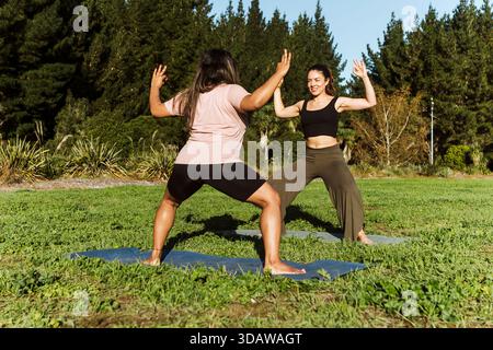 Two women practicing yoga together outdoors on a grassy field in New Zealand during autumn. They are in a calming environment surrounded by lush green Stock Photo