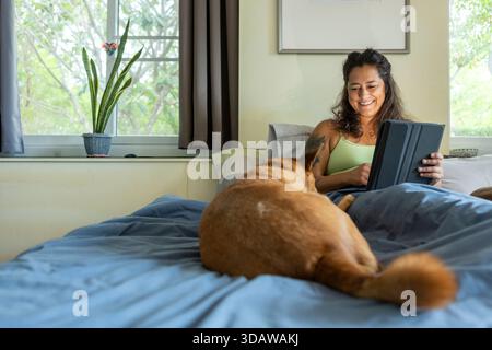 Woman comfortably seated in bed, smiling while using a tablet. A dog rests by her side, creating a cozy and relaxed atmosphere. Natural light illumina Stock Photo