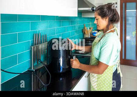 Side view of a woman stands in her kitchen, wearing a green apron, using an air fryer to prepare homemade bread. She carefully adjusts the settings, s Stock Photo