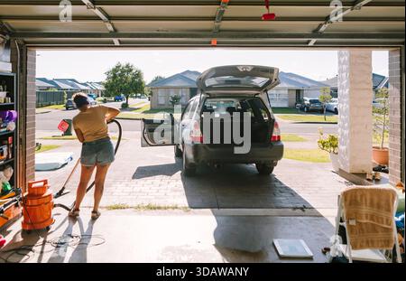A woman is busy cleaning her car in the driveway of her suburban home in Australia. The scene captures a sunny day as she vacuums and tidies the vehic Stock Photo