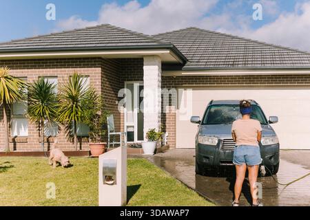 A woman washes her car in the driveway of her modern home in Australia. Surrounded by greenery and bright sunshine, she focuses on maintaining and cle Stock Photo