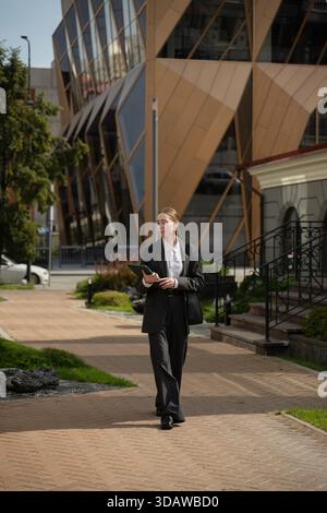 A business woman in a suit walks confidently through a bustling cityscape, holding a digital tablet. The modern architecture and greenery complement t Stock Photo