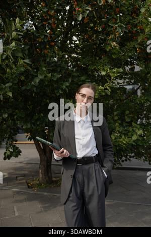 Professional business woman in a smart suit stands confidently in an urban environment, holding a tablet. The city backdrop adds a sense of dynamism a Stock Photo