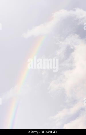 A delicate rainbow arches through a cloudy sky, blending soft pastel colors with the serene hues of the clouds. The gentle gradient creates a captivat Stock Photo