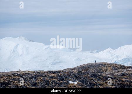 Back view of unrecognizable hikers standing on a hill overlooking vast, icy landscapes in Ilulissat Icefjord, Greenland. Majestic snow-covered mountai Stock Photo