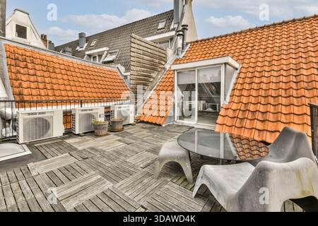 An urban rooftop terrace featuring modern chairs and a round glass table is surrounded by red-tiled roofing. Air conditioning units add a functional a Stock Photo