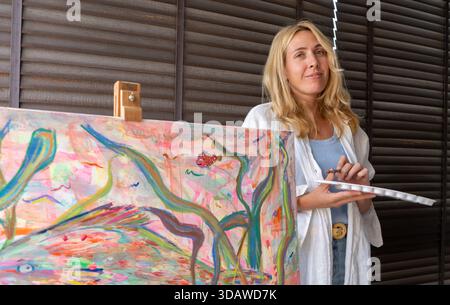 A woman artist stands beside her colorful canvas, holding a palette and brush. She is painting an abstract design with vivid colors, capturing creativ Stock Photo