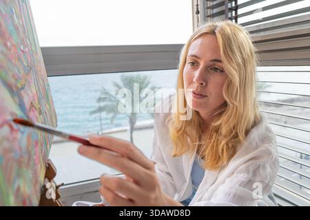 A focused woman artist skillfully painting on a canvas by the sea. She captures the essence of the ocean view outside her window, blending colors with Stock Photo