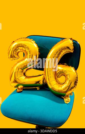 Festive golden number balloons form the number 26, placed on a blue chair with a vibrant yellow background, perfect for celebrating birthdays or anniv Stock Photo
