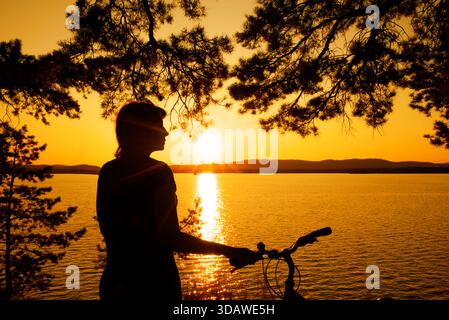 A tranquil scene of an individual with a bicycle, silhouetted against a stunning sunset over a serene lake. Framed by tree branches, it captures the e Stock Photo