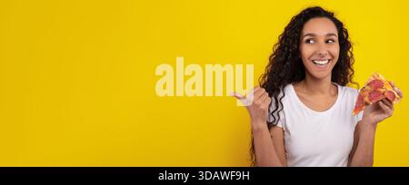 Young Woman Enjoys Pizza Against a Bright Yellow Background While Smiling and Pointing Stock Photo