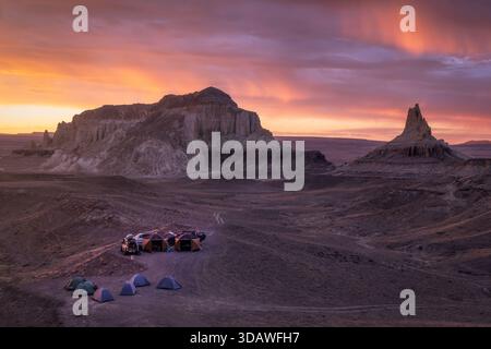 View of tents clustered on the arid ground under a vibrant sunset sky, shadowed by towering rock formations, Aktau, Mangystau Region, Kazakhstan. Stock Photo