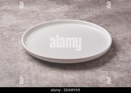 empty matte white ceramic plate with round rim on textured grey concrete stone background surface, rustic dining or menu concept with copy space Stock Photo