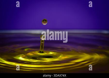 Vibrant yellow water splash with suspended droplet on purple background, creating dynamic abstract liquid art with bright reflections. Stock Photo