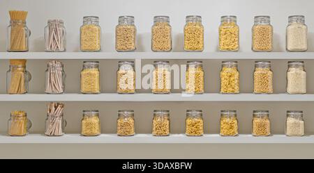 A shelf full of glass jars with different colored pasta. The jars are arranged in rows, with some jars containing spaghetti and others containing penn Stock Photo
