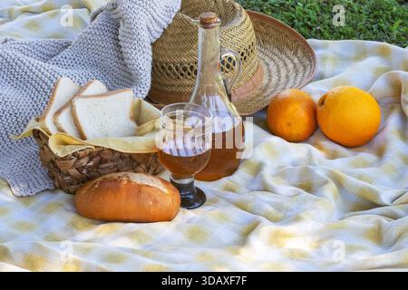 Picnic spread on yellow white plaid blanket with sliced and round bread, wicker basket, jug and glass of juice, two oranges and straw hatsunny, rustic Stock Photo