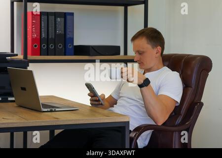 Multitasking professional viewing phone notifications with coffee in hand beside laptop in modern office setup. (96 characters) Stock Photo