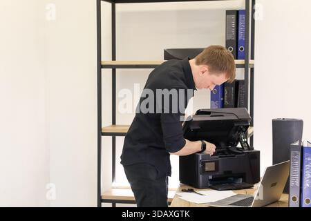 Man in office leans in to closely observe opened printer for issues, surrounded by binders and laptop on desk. Stock Photo