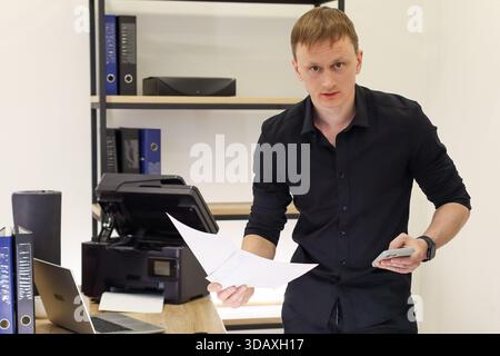 Caucasian male in black shirt holds paper and smartphone while standing in office near printer and desk. Stock Photo